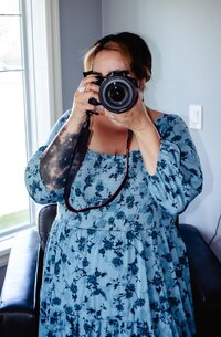Woman in a blue floral, flowy dress holding a professional camera in front of her face.