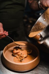 Close-up of sauce being spooned over a seafood dish with prawns in a tan ceramic bowl.