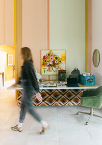 Motion-blurred branding photo of a woman walking past a vibrant floral painting and art-filled desk in a retro workspace, shot in Greenville SC by Dani Lacey Photographs.