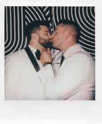A polaroid of two men in white shirts and suspenders sharing a kiss