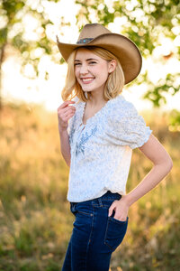 A Littleton senior girl holds her blonde hair while wearing a cowboy hat and smiling at a Littleton Senior photographer