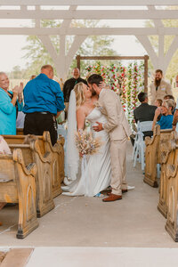 A newly married couple shares a kiss down the aisle in a rustic open-air wedding chapel at Legacy Farm in Stonefort, Illinois, surrounded by guests and wooden pews.
