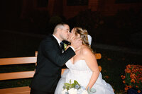 A bride and groom share a kiss while sitting on a bench at night. Captured by an NJ wedding photographer, the bride holds a bouquet in her white dress, while the groom, in a black suit, makes the moment timeless.