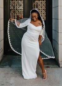 An African American bride poses while holding her veil in both hands in front of an ornately decorated black door on a white stone building. She's wearing a long sleeved white dress and smiling.