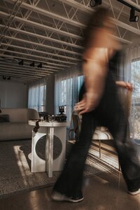 A blurred, motion-filled image captures Andrea Cakmar walking across a room filled with natural light. A table with a camera and photography equipment is visible in the background.