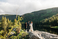 Couple getting married with an officiant on a rock. There's a lake, a mountain and trees behind them