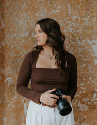 Photographer looking over her shoulder in a brown top holding a camera in her hands.