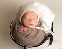 Newborn in a bucket with a white bonnet
