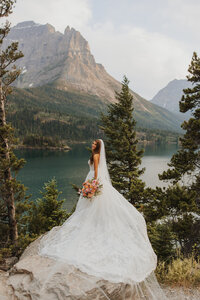 Bride smiling at the camera while standing on a rock overlooking a lake, with trees and mountains behind her in Glacier National Park.