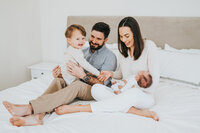 Family of four sits and laughs together on white bed during in-home newborn photography session