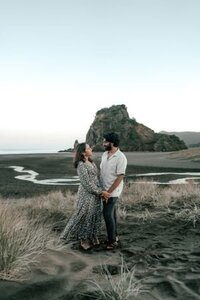 Keerthana and Suraj walking hand in hand through the black sand beach dunes with dramatic cliffs at blue hour during their Auckland engagement session, captured by Zanthe Vorsatz.