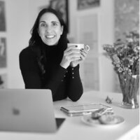 woman sitting at a desk with a table and laptop in front of her holding a cup of coffee