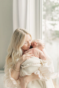 Mother gently snuggles her newborn on her shoulder, standing in natural light during a soft, quiet in-home newborn session in Kansas City.