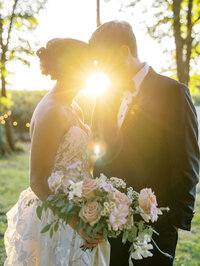 A bride and groom stand close to each other with sunlight shining through, representing the photography style of Among the Oaks. 