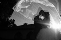 Black and white wedding photograph showing a bride and groom in silhouette embracing under a flowing wedding veil at the White Room in historic downtown St. Augustine. The dramatic lighting creates a romantic backlit effect with the veil catching the light beautifully against the building's architecture.