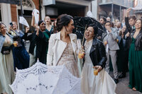 Two brides in wedding attire, holding drinks and parasols, smiling and walking through a crowd of guests at Hotel Mazarin in New Orleans.