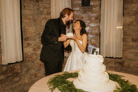 A newlywed couple laughing as they hold a piece of wedding cake 