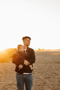 A father holds his baby while the sun sets behind them during their family photoshoot at a local christchurch beach with photographer lucy jane