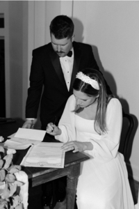 The bride is sitting and signing the marriage certificate while the groom is standing.