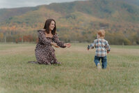 A mother squats in a field with her hands out for her young son to run to her. They are in front of a large mountain with fall colors. 