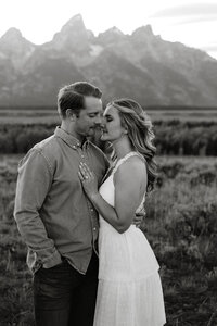 man and woman embracing while standing in a field with mountains in the background