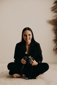 Smiling Mullingar photographer in a black outfit sitting cross-legged and holding her camera