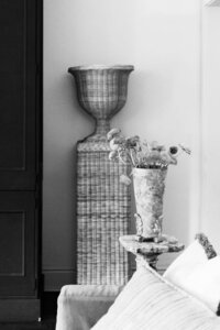 Black and white photo of a living room corner styled by Sweet Pea Ink Creative, featuring a vase of flowers on a small table, a woven decorative piece, and part of a couch with cushions.