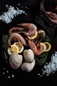 A rich and dramatic image of a seafood platter featuring shrimp, clams, lemon slices, and fresh herbs, presented on a dark background. Captured by Mica McCook, this photograph emphasizes the freshness and elegance of a gourmet seafood dish.