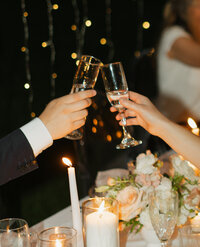 The bride and groom clinking champagne glasses during their reception. The photo is cropped so you only see their hands and part of their arms.