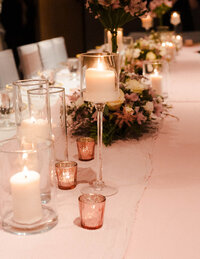 A detailed view of a wedding table with glowing candlelight from glass holders, surrounded by pink and white floral arrangements on a soft pink table runner.