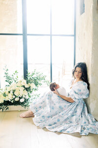 a mother sits in a big window with beautiful light, wearing a stunning blue dress and holding her newborn on her lap. photo taken by San Antonio Newborn Photographer Teressa Jane