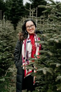 Woman standing among evergreen trees, smiling warmly in a red turtleneck sweater and colorful patterned scarf on a cool winter day.
