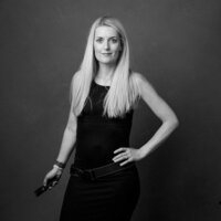 Black and white self-portrait of photographer Katie Campbell, standing confidently with one hand on her hip and the other holding a camera remote. She wears a fitted black dress with a belted waist, her long blonde hair framing a poised and self-assured expression. A powerful, unfiltered moment from 'THIS IS ME – A Photography Experience by Katie Campbell' in Atlanta, GA, celebrating self-expression and the artist behind the lens.
