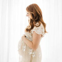 Elegant side profile of an expecting mother in a delicate embroidered gown, gently holding her baby bump near soft curtains.
