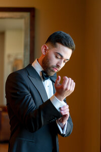 A groom in a tuxedo is adjusting his tie, showcasing a polished and sophisticated appearance at his wedding.