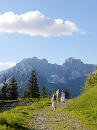 Familie spaziert auf einem Bergpfad mit Blick auf die Alpen im warmen Abendlicht.