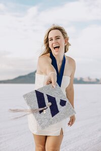 Girl in white dress smiling holding a Westminster University graduation cap for senior portraits at the Salt Flats in Utah 