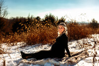 A woman is sitting on a fuzzy blanket. She's pregnant with a big bump and holding the bump with one hand and one hand behind her. She's in a black dress. There's snow on the ground and tall brown grass behind her.