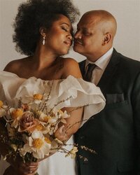 A couple shares an intimate moment, gently leaning their heads together. One person, wearing a white off-the-shoulder outfit, holds a bouquet of flowers, while the other, dressed in a dark suit, gazes at them lovingly.