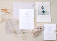 A wedding invitation suite flat lay with an engagement ring and wedding band on a round pink ring box