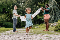 A young child in a light blue dress runs happily toward the camera while two adults hold a blanket behind them, creating a playful atmosphere in a park setting.