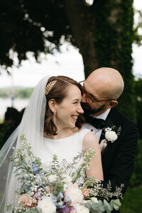 Groom holding his bride while she looks off into the river laughing. She's holding florals with pastel colors and is wearing a chic dress and veil. 