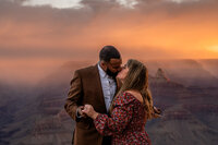 A couple stands together on the edge of the Grand Canyon, with the sun setting in the background as the man proposes, his partner in awe of the breathtaking view.