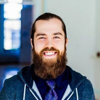 This is a portrait of a man with a friendly smile, looking directly at the camera. He has a full beard and long hair tied back. He is wearing a blue hoodie and a shirt with a tie underneath. The background is bright, with windows that suggest the photo was taken indoors during daylight. The man's appearance and the setting give the photo a casual yet professional feel.