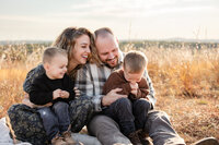 a family of 4 hug eachother while sitting in a grassy field at sunset. The mom and dad are laughing at the two boy children, as they all wear fall sweaters and smile at eachother.
