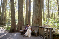 Bride and groom sit on a wooden bench in a serene cedar forest during their Montana elopement. The bride wears a delicate embroidered wedding dress, leaning against the groom, who is dressed in a burgundy suit. Sunlight filters through the tall trees, creating a peaceful and intimate moment in nature.