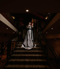 Dramatic wedding portrait of a bride in a black-trimmed gown and groom in dark attire, embracing on a grand staircase under moody lighting, captured with a cinematic, gothic-inspired style by Auburn Blue Photography.