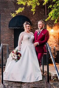 Bride holding a bouquet and groom wearing a burgundy suit while posing on a bridge.