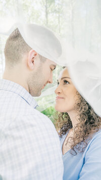 A bride and groom gaze into each other's eyes under a wedding veil at their engagement shoot