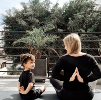 A moment of connection is captured between an adult and a child, both sitting back-to-back in a peaceful, mirrored pose that suggests mutual support and harmony.